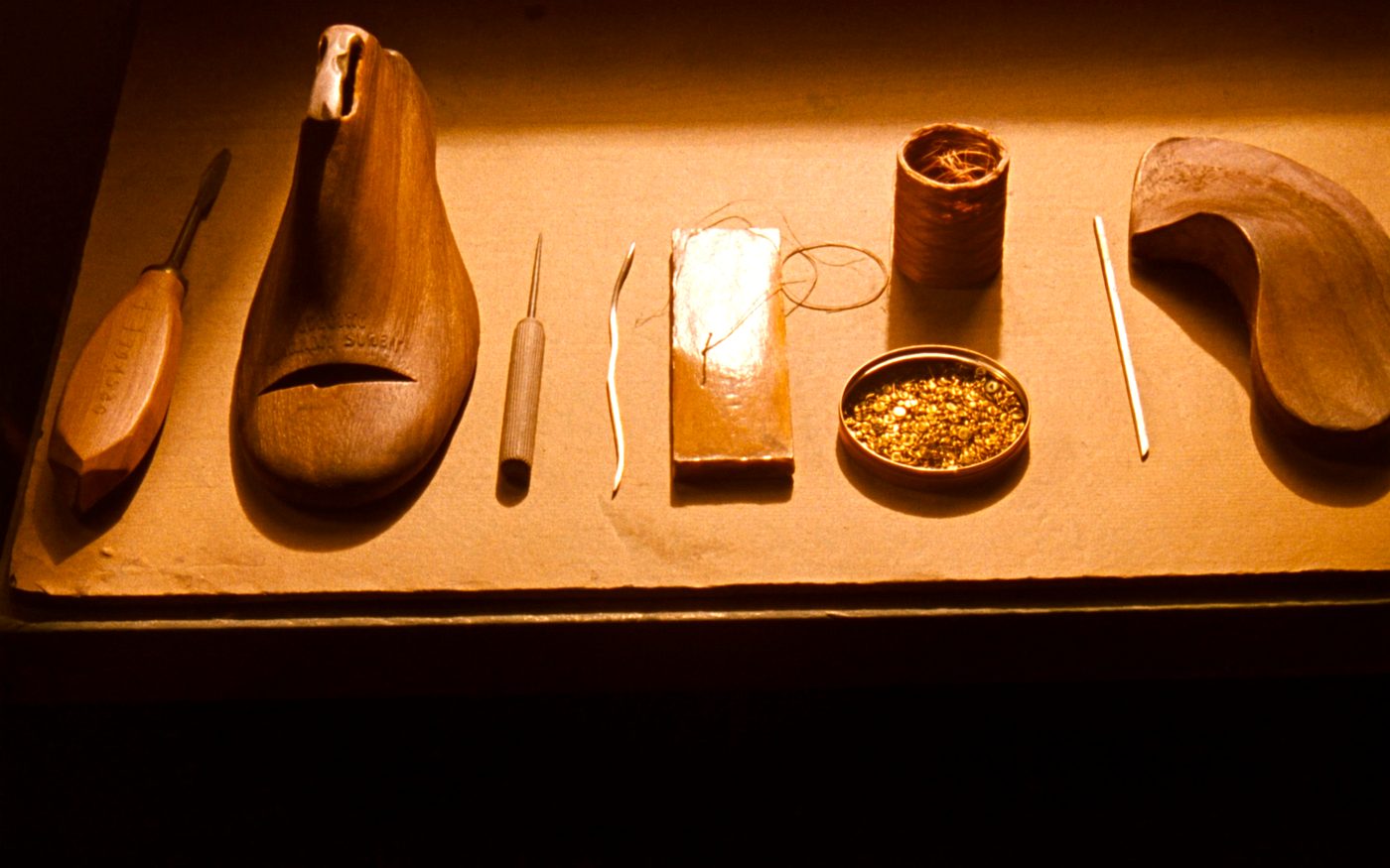 A wooden shoe last, a knife, an awl, a curved needle threaded with waxed linen, a block of wax, a spool of thread and a tin of brass tacks laid out in a row on brown paper.