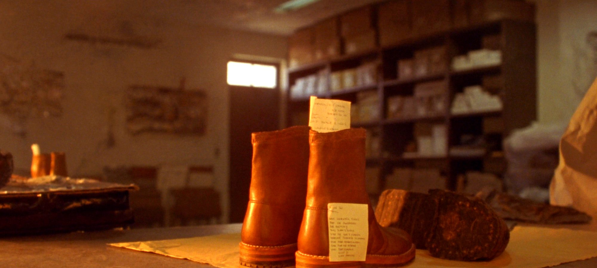 A pair of tan leather boots standing on kraft paper on the shop counter, paper repair tickets tucked into them, shelves of boxed shoes behind.