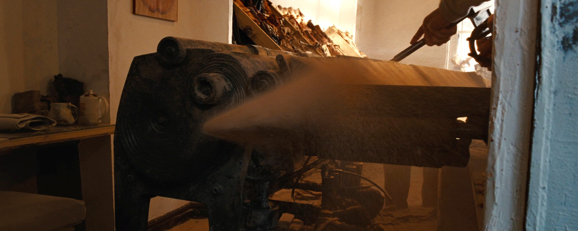A cast-iron bench machine running in the workshop, throwing a fine plume of dust across the light, offcuts of leather stacked behind it.