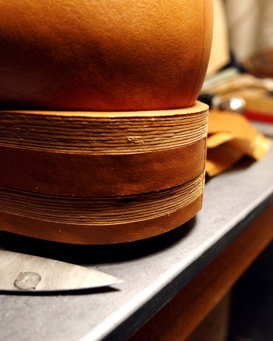 A heel block on a steel bench, built up from stacked layers of leather with the trimmed edges showing, a trimming knife lying in front of it.