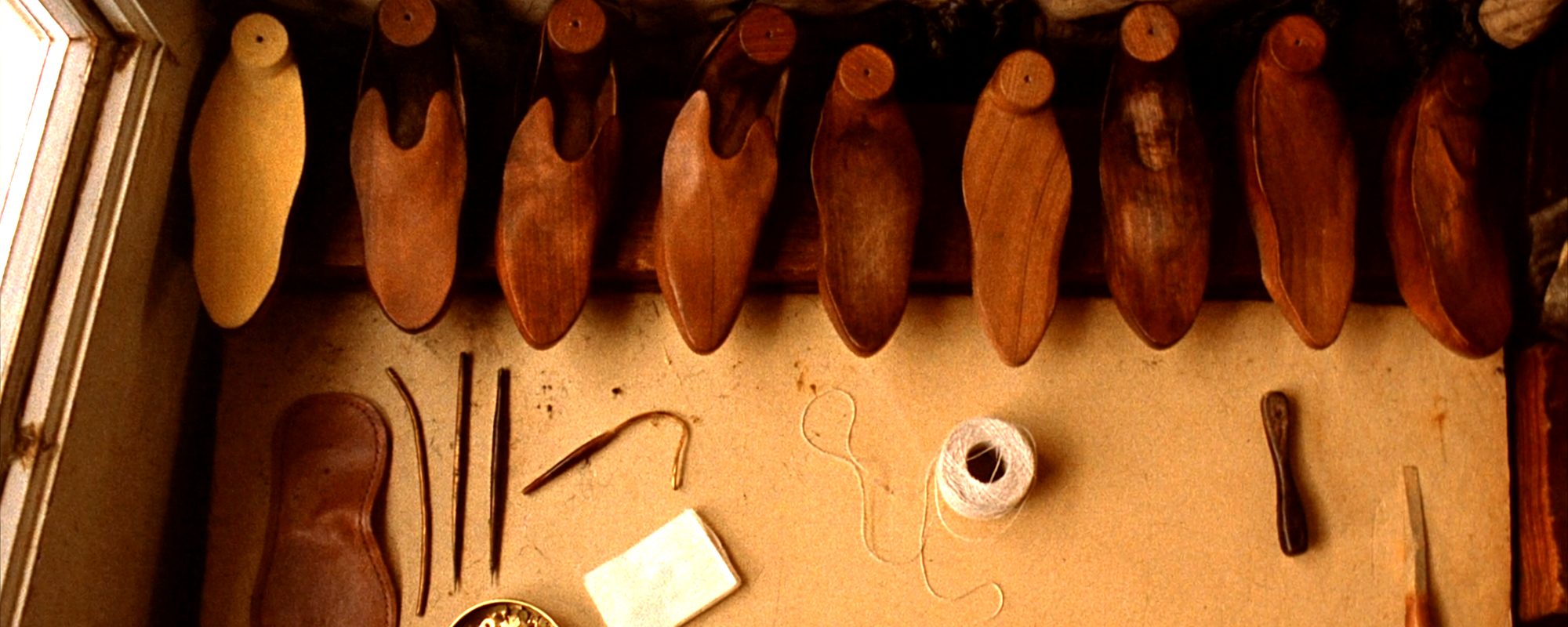 A row of wooden shoe lasts hanging above a workbench, with a cut leather half sole, awls, a curved needle, waxed thread, a tin of tacks and a steel shank laid out below them.