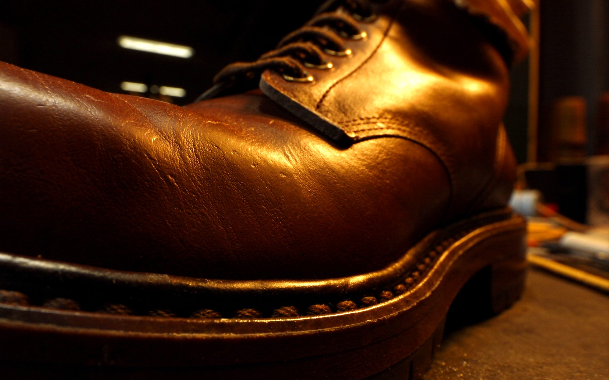 A well polished brown leather work boot on the bench, warm light raking across the vamp and the brass speed hooks, the stitched sole edge in shadow underneath.